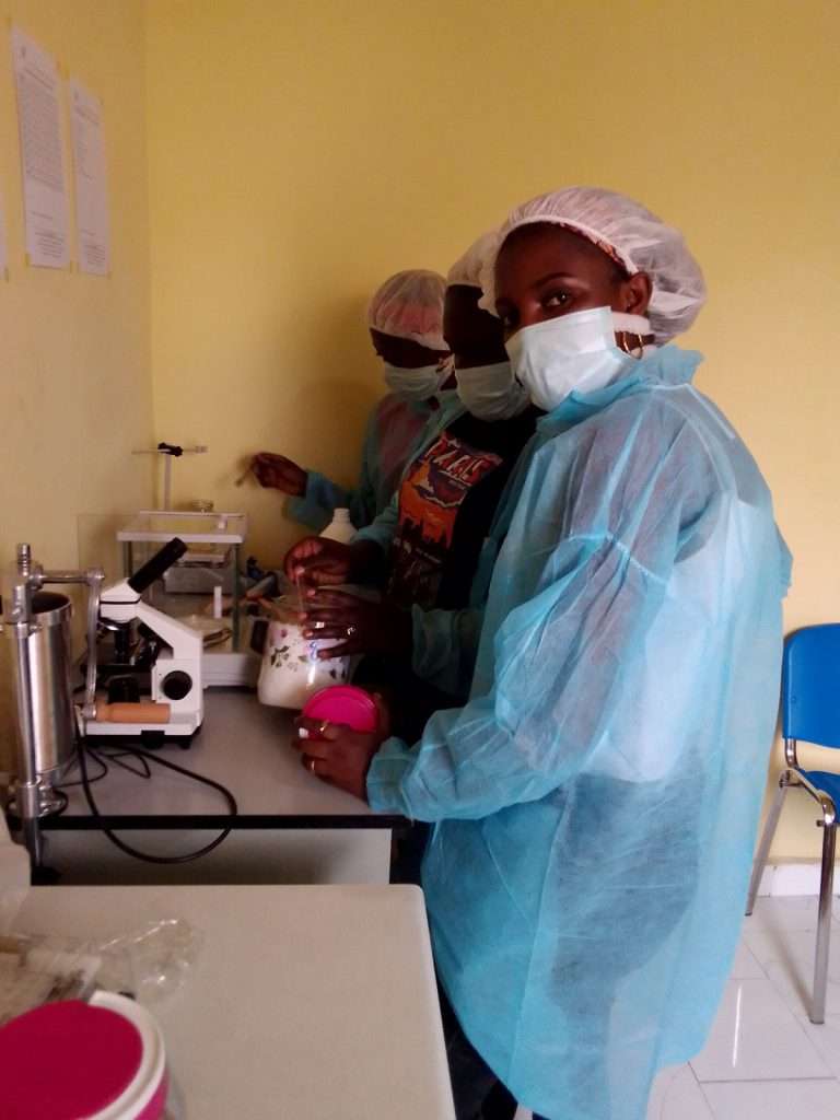 In un laboratorio in Congo dedicato alla produzione e allo sviluppo di spirulina per combattere la malnutrizione infantile, tre operatrici lavorano a un banco da laboratorio. La donna in primo piano, che guarda l'obiettivo, indossa un berretto chirurgico bianco, una maschera azzurra e un camice protettivo azzurro. Tiene in mano dei contenitori con polvere bianca (spirulina), tra cui un barattolo con coperchio rosa. Dietro di lei, le sue colleghe, anch'esse con DPI (berretti, maschere, camici), sono impegnate a misurare e lavorare la polvere con l'ausilio di bilance da laboratorio, un microscopio bianco, un estrusore manuale e vari barattoli di vetro. L'ambiente è pulito, con pareti gialle pallide sulle quali sono appesi dei documenti. L'immagine cattura un momento di lavoro pratico e professionale.