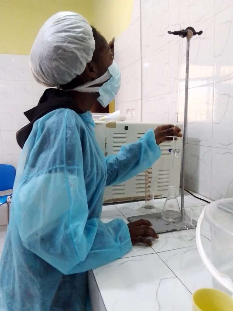 In un laboratorio chimico, una giovane donna indossa un camice monouso azzurro, una retina copricapo bianca e una mascherina chirurgica mentre esegue una procedura di titolazione. È in piedi davanti a un bancone piastrellato in bianco e sta manipolando con la mano destra il rubinetto di una buretta fissata a un supporto. Sotto la buretta ci sono un cilindro graduato e una beuta. Sullo sfondo si nota un incubatore.