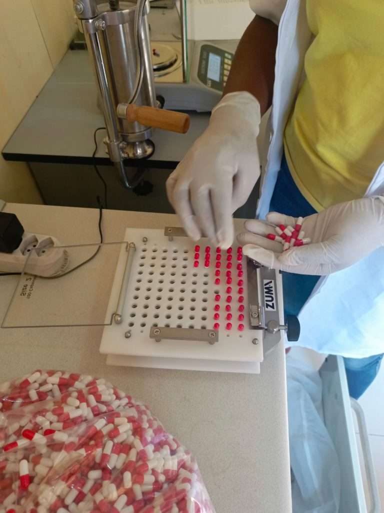 In un dettaglio ravvicinato, un operatore di laboratorio con guanti bianchi e camice sta utilizzando una macchina incapsulatrice manuale per confezionare gli integratori.
Le mani inseriscono con precisione delle capsule bicolore (bianche e rosa/rosso) nei fori di una piastra bianca per essere riempite con la polvere di spirulina. In primo piano spicca un grande sacchetto trasparente colmo di capsule già pronte per la distribuzione. Sullo sfondo del banco da lavoro si notano una bilancia elettronica di precisione e un estrusore manuale in metallo, strumenti essenziali per garantire il corretto dosaggio del supplemento nutrizionale.